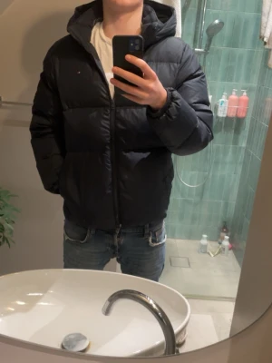 Tommy Hilfiger jacka  - Mötkblå pufferjacka från Tommy Hilfiger med huva och diskret logga på bröstet. Jackan har dragkedja framtill och är quiltad för extra värme. Perfekt för kalla dagar och har en clean, modern look som funkar till jeans och sneakers. STROLEK S/176