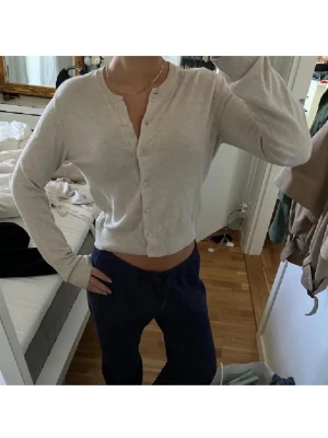 Beige kofta med knappar - Säljer en beige, croppad kofta med rund hals och långa ärmar. Koftan har knappar hela vägen framtill och är gjord i ett mjukt stickat material som känns skönt mot huden. Perfekt att slänga på över ett linne eller t-shirt för en chill look.
