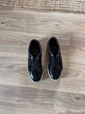 Svarta sneakers med lackad tå - Snygga svarta sneakers med snörning och rund tå. Skorna har en cool lackad detalj på tån och ovandel i textil. Perfekta för dig som gillar stilrena och moderna skor med en twist.