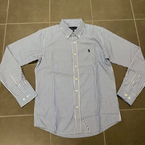 Blårandig skjorta från Ralph Lauren stl M - Klassisk blårandig skjorta från Ralph Lauren i bomull med button-down krage och broderad logga på bröstet. Skjortan har lång ärm, vita knappar och en relaxed classic fit. Perfekt till jeans eller chinos för en clean look.