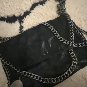 Svart axelväska med kedjedetaljer - Snygg svart axelväska med glansig finish och kraftiga silverfärgade kedjor som axelrem och runt kanterna. 