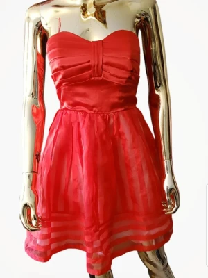 Röd axelbandslös kortklänning från Vila - Säljer en röd axelbandslös kortklänning från Vila i modellen Celian Dress. Klänningen har ett plisserat liv, markerad midja och ett skirt, randigt tyg i kjolen. Materialet är glansigt och ger en lyxig känsla. Perfekt för dig som vill sticka ut med färg och stil.