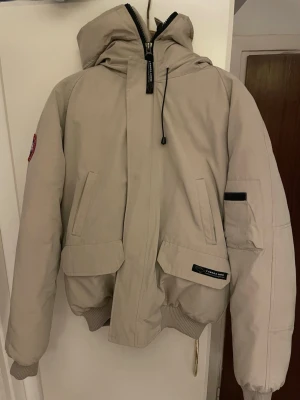 Beige dunjacka från Canada Goose - Säljer en beige dunjacka från Canada Goose med huva och ribbade muddar. Jackan har flera fickor, dragkedja och patch på ärmen. Insidan är svart och fodrad med dun för extra värme. Perfekt för kalla dagar och har en clean, stilren look. Storleken är XXL, men den är liten för att vara det, skulle säga att den passar bättre som L för en man och L/XL för en kvinna. 