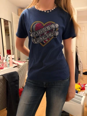 Blå t-shirt med hjärtmotiv - Säljer en zadig voltaire blå t-shirt med korta ärmar och ett stort hjärtmotiv framtill i rött, vitt och gult. Texten på hjärtat är dekorerad med små glittriga detaljer. Skön passform och tillverkad i mjuk bomull, perfekt till jeans.