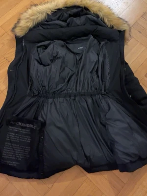 Svart Rockandble dunjacka med päls - Svart dunjacka från Rockandble med fluffig fuskpäls på huvan. Jackan har flera dragkedjeförsedda fickor, knappar framtill och snörning i midjan för snygg passform. Perfekt för kalla dagar med sin långa modell och varma material.