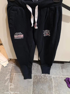 Svarta mjukisbyxor Vinson Polo Club S - Svarta mjukisbyxor från Vinson Polo Club i storlek S. Byxorna har vita snören i midjan, broderade loggor och amerikansk flagga på framsidan. Tillverkade i mjukt bomullsmaterial med ribbade muddar vid benslut. Perfekta för chill eller träning.