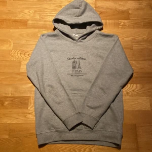 Hoodie från studie silvano - Grå hoodie från Studio Milano med tryck av Eiffeltornet och texten 'Paris' på bröstet. Klassisk huva och magficka, perfekt för en chill och avslappnad stil. Mjuk och skön bomullsblandning, långärmad modell.