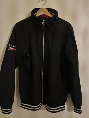 Svart bomberjacka från E.T.R.L - Svart bomberjacka med vita ränder på muddar och nederkant. Jackan har dragkedja framtill, hög krage och broderad logga på ärmen med en liten flagga. Snygg och sportig stil som passar till många outfits.