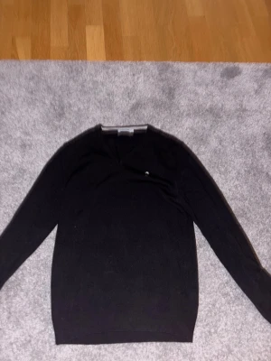 Svart v-ringad tröja från J Lindeberg  - Svart v-ringad tröja från J Lindeberg med diskret logga på bröstet. Tröjan är långärmad och har en klassisk, stilren look som passar perfekt till jeans eller chinos. Tillverkad i mjukt material som känns skönt mot huden.