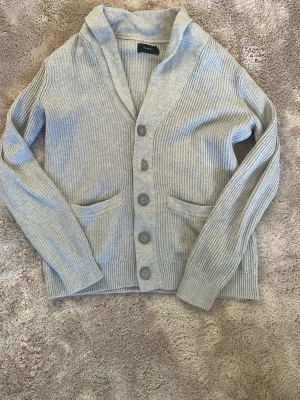 Grå stickad kofta från Reserved - Säljer en grå stickad kofta från Reserved med ribbad struktur och stora knappar framtill. Koftan har två fickor och en bred krage som ger en chill vibe. Perfekt att slänga på sig när det är lite kyligt ute.