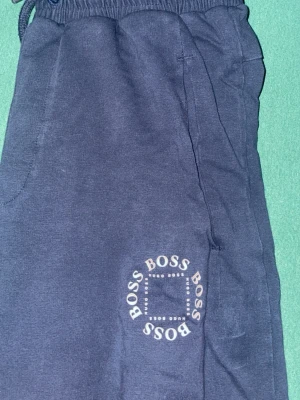 Mörkblå mjukisbyxor från Hugo Boss - Snygga mörkblå mjukisbyxor från Hugo Boss med tryckt logga i vitt på benet. Byxorna har elastisk midja med snörning och är gjorda i mjukt bomullsmaterial. Perfekta för chill eller träning, med normal passform och praktiska sidofickor.