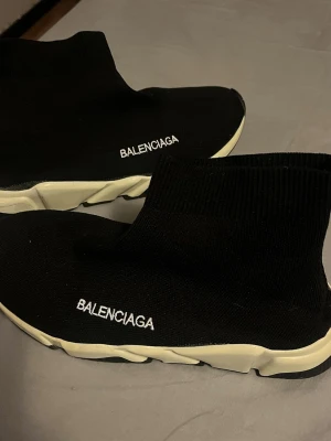 svarta sock sneakers - Svarta sock sneakers med vit sula och broderad logga på sidan. Skorna har en stickad ovandel i syntetmaterial som sitter tight runt foten och en chunky, platt sula i vitt. Modern och clean design som är riktigt populär just nu. Passar både man och kvinna, storlek 39. Pull on modell.