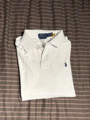 Vit piké från Polo Ralph Lauren - Vit klassisk pikétröja från Polo Ralph Lauren med korta ärmar och krage. Tröjan har två knappar vid halsen och den ikoniska blå broderade loggan på bröstet. Materialet är mjuk bomull som känns skönt mot huden. Perfekt för en clean och stilren look.