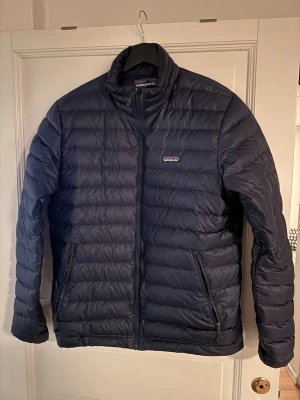 Mörkblå dunjacka från Patagonia - Snygg mörkblå dunjacka från Patagonia med quiltad design och dragkedja framtill. Jackan har två sidofickor med dragkedja och en klassisk ståkrage. Perfekt för kyliga dagar och har en clean look som funkar till det mesta.