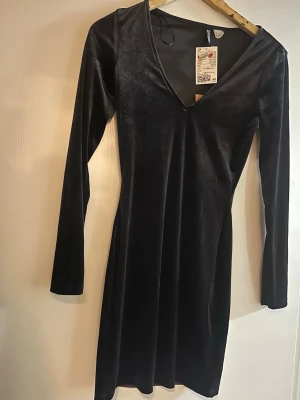 Svart sammetsklänning från H&M - Snygg svart klänning i mjuk sammet från H&M med lång ärm och v-ringad hals. Klänningen har en figurnära passform och är knälång, perfekt för dig som gillar stilrena och eleganta plagg. Enkel design utan mönster eller extra detaljer.