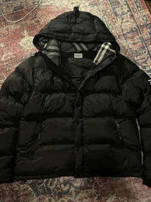 Svart pufferjacka från Burberry - Svart pufferjacka från Burberry med huva och klassiskt rutigt foder. Jackan har dragkedja och knappar framtill, samt två sidofickor med dragkedja. Märkespatch på ärmen och quiltad design. Perfekt för kalla dagar.