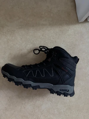 Svarta vinterkängor från Lanner - Svarta kängor från Lanner med grov sula och snörning. Ovandelen är i slitstarkt syntetmaterial och skinn, fodrade med mjukt tyg för extra värme. Perfekta för kalla dagar och har en robust design med detaljer i grått och vita sömmar.