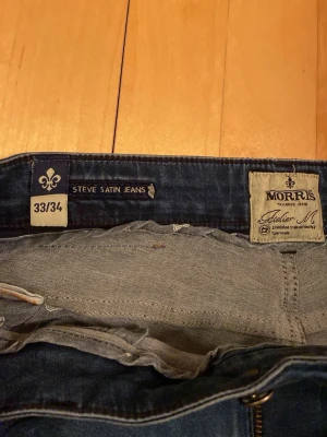 Morris Steve Satin Jeans blå 33/34 - Blå jeans från Morris, modell Steve Satin Jeans. Klassisk femficksdesign med snygga detaljer och mörkblå tvätt. Märkespatch i brunt skinn baktill och diskret broderad bakficka. Materialet är jeans med en mjuk känsla.
