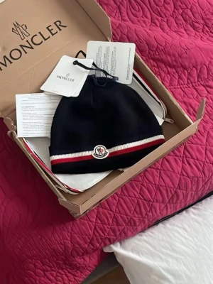 Svart Moncler mössa med röd/vit kant - Snygg svart mössa från Moncler med klassisk logga framtill och en stilren kant i rött och vitt. Perfekt accessoar för att hålla värmen och samtidigt se riktigt fräsch ut. Mössan levereras i originalförpackning med tags kvar.