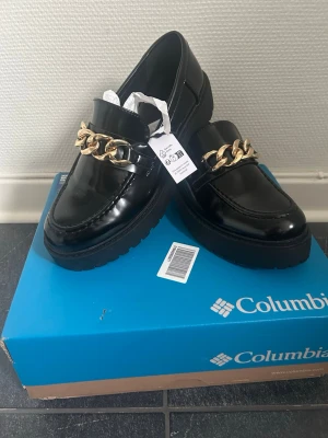 Svarta loafers med guldkedja Even&Odd - Snygga svarta loafers från Even&Odd med chunky guldkedja framtill. Skorna har en rund tå och platt sula, tillverkade i blankt syntetmaterial som ger en cool och modern look. Perfekta att lyfta din outfit med lite extra edge.