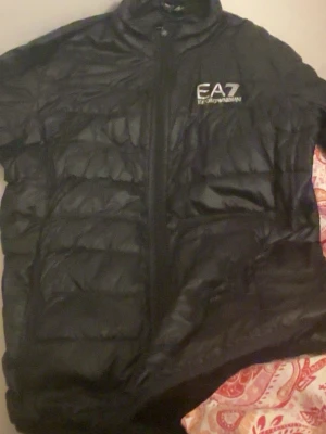 Svart pufferjacka från EA7 Emporio Armani - Svart pufferjacka från EA7 Emporio Armani med glansig yta och diskret logga på bröstet. Jackan har ståkrage, dragkedja framtill och quiltad design. Perfekt för dig som vill ha en snygg och lätt jacka med sportig vibe.