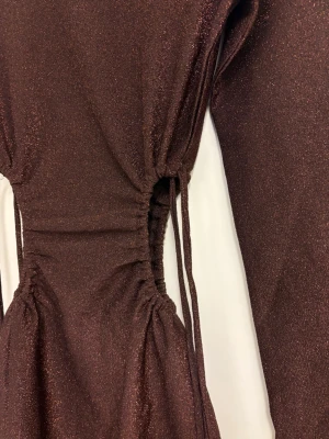 Glittrig brun klänning med snörning - Snygg brun glittrig klänning från hm med långa ärmar och snörning i midjan som ger en cool siluett. Kort modell med slits framtill och rund halsringning. Perfekt för dig som vill sticka ut med lite extra glitter.