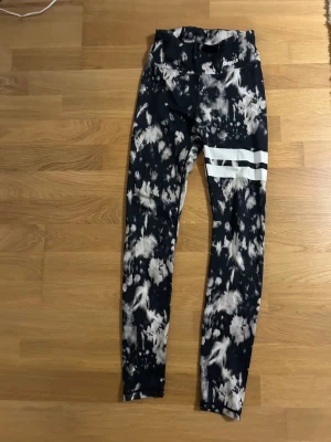 Svarta och vita leggings från Stronger - Snygga leggings från Stronger i storlek XS med svart och vit batikmönster och två vita ränder på ena benet. Hög midja och stretchigt material för skön passform. Säljer då den ej används längre