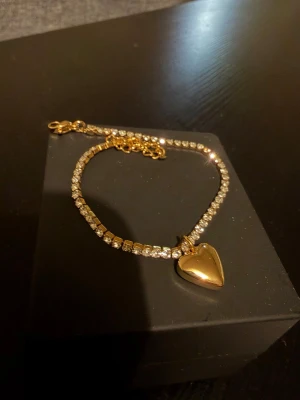 Tennis armband med berlock - Ett vackert 18k gult Guldpläterad armband med en hjärtformad berlock. Armbandet är dekorerat med strass stenar av bästa zirconia kvalitet.   Anti Rostfritt går att bada och duscha med!   Tjocklek 2mm längd 17cm + 5cm om det behövs!   Kommer i en liten smycken påse! 