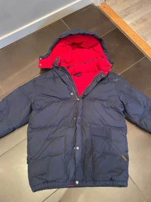 Mörkblå pufferjacka från Polo Ralph Lauren - Säljer en mörkblå pufferjacka från Polo Ralph Lauren med röd insida och huva. Jackan har dragkedja, knappar och två stora fickor med mjukt foder. Den är i storleken XL med funkar som M. Perfekta vinterjackan!