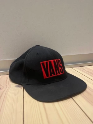 Svart Vans keps med röd logga - Säljer en svart keps från Vans med stor röd broderad logga framtill och texten 'Since 1966' baktill. Kepsen har böjd skärm och justerbar snapback för perfekt passform. Perfekt för dig som gillar streetwear och vill ha en snygg accessoar.