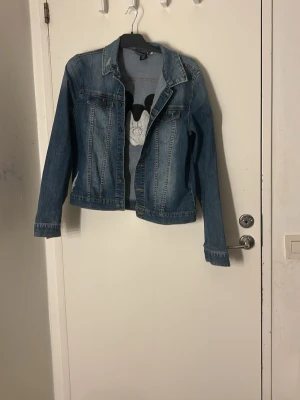 Jeansjacka med Mickey Mouse  - Säljer en blå jeansjacka med broderad Mickey Mouse på ryggen och en patch med texten 'Mickey Mouse' nedtill. Jackan har klassisk krage, knappar framtill och två bröstfickor. Perfekt för dig som vill sticka ut med en ikonisk Disney-detalj. Pris går att diskutera Tvätt innan leverans 