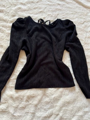 Svart långärmad blus med snörning - Säljer en svart blus med lång ärm och puffiga axlar. Toppen har ribbad struktur och en snygg öppen rygg med snörning i nacken. Materialet känns mjukt och stretchigt, perfekt för dig som gillar detaljer och en cool siluett.