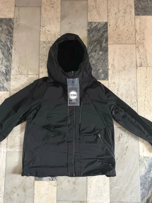 Svart vindjacka från Colmar Revolution - Svart vindjacka från Colmar Revolution med huva och flera dragkedjor framtill. Jackan har en clean look med snygga linjer och praktiska fickor med dragkedja. Perfekt för dig som gillar stilren och funktionell design.