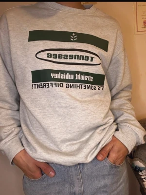 Grå sweatshirt med Tennessee-tryck - Säljer en grå sweatshirt med grönt och svart Tennessee-tryck på bröstet. Tröjan har rund hals och långa ärmar med ribbade muddar. Perfekt för dig som gillar casual och streetwear. Passar till jeans eller joggers.