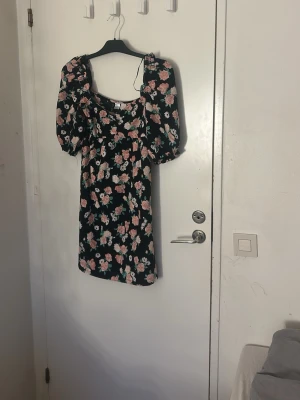 Blommig kortklänning från H&M - Supersöt svart kortklänning från Divided med puffiga kortärmar och ett färgglatt blommigt mönster i rosa och vitt. Klänningen har en rundad hals och är gjord i mjuk bomull, perfekt för dig som gillar romantisk stil och vill sticka ut, Pris kan diskuteras och tvätt ingår innan leverans 