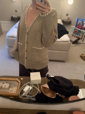 Beige kofta från Sandro med vita detaljer - Superfin beige kofta från Sandro med vita tvinnade detaljer längs kanterna och fickorna. Koftan har v-ringning, knappar framtill och två stora fickor. Perfekt för lager-på-lager och chill vibes. Storlek 1 ifrån Sandro! Helt ny knappt använd.