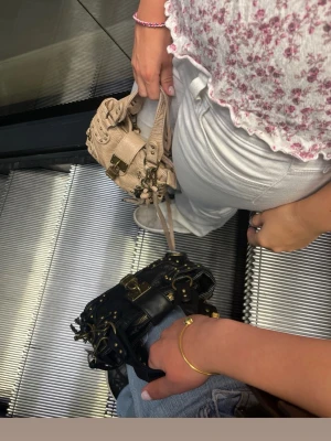 Svart handväska med nitar och spännen - Säljer en svart handväska med coola guldfärgade nitar och detaljer. Väskan har ett snyggt spänne framtill och flera metalldetaljer som ger den en edgy vibe. Perfekt storlek för det viktigaste och har en kort rem för att bära i handen.