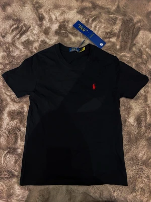 Svart t-shirt från Polo Ralph Lauren - Svart t-shirt från Polo Ralph Lauren med klassisk rund hals och korta ärmar. På bröstet finns den ikoniska röda Polo-loggan broderad. T-shirten är gjord i mjuk bomull och har en clean, stilren look som funkar till allt.