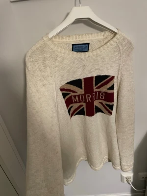 Vit stickad tröja Morris  - Säljer en vit stickad tröja från Morris Co i storlek medium. Tröjan har ett stort Union Jack-motiv med texten 'Morris' på bröstet. Rund hals och långa ärmar, perfekt för dig som gillar klassisk brittisk stil. Materialet är mjukt och skönt stickat.