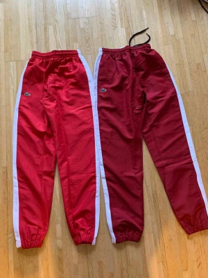 Röda träningsbyxor från Lacoste - Två par röda träningsbyxor från Lacoste med vita sidopaneler och elastisk midja. Klassisk Lacoste-logga på benet och avslut med resår vid anklarna. Perfekta för sport eller chill. Ena paret har snörning i midjan.