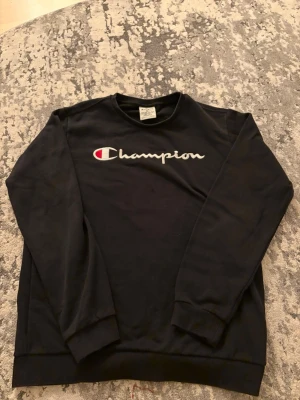 Svart sweatshirt från Champion XL - Svart sweatshirt från Champion i storlek XL. Tröjan har lång ärm, rund hals och ett broderat Champion-logo på bröstet samt en liten logga på ärmen. Tillverkad i mjuk bomull och polyesterblandning, perfekt för chill eller streetwear.