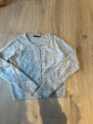 Grå kofta från 157, storlek S - Mysig grå kofta från 157 med rund hals och knappar framtill. Koftan är långärmad och har en ribbad kant vid hals, ärmslut och nederkant. Perfekt att slänga på sig när det är lite kyligt ute. Materialet känns mjukt och stickat. Strl S