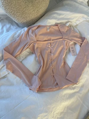 Ljusrosa långärmad topp från Nelly - Supersöt ljusrosa långärmad topp från Nelly i mjuk bomull. Toppen har en figurnära passform och dekorativa rosetter framtill som ger en gullig touch. Perfekt för dig som gillar romantiska detaljer och vill ha något som sticker ut lite extra. 💕 