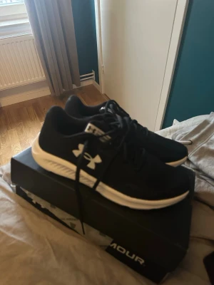 Svarta Under Armour sneakers - Säljer ett par svarta sneakers från Under Armour med vit sula och klassisk logga på sidan. Skorna har snörning och är tillverkade i ett syntetmaterial med mesh för bra ventilation. Perfekta för träning eller chill vardagsstil.