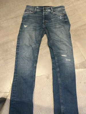 Blå jeans med slitningar - Säljer ett par klassiska blå jeans från Jack and Jones  med snygga slitningar och lätt tvättad look. Modellen har raka ben och fem fickor, samt dragkedja och knapp i midjan. Perfekt för dig som gillar en avslappnad stil med lite edge.