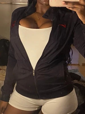 Svart Nike vindjacka PSG - Svart vindjacka från Nike med PSG-logga på bröstet och röd swoosh. Jackan har dragkedja framtill, ståkrage och två sidofickor. Materialet är lätt och känns sportigt, perfekt för chill eller när du är på språng. Snygg och enkel design med långärmade ärmar.