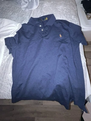 Blå pikétröja från Polo Ralph Lauren - Säljer en klassisk blå pikétröja från Polo Ralph Lauren med korta ärmar och krage. Tröjan har tre knappar framtill och den ikoniska broderade loggan på bröstet. Perfekt för en chill och stilren look.