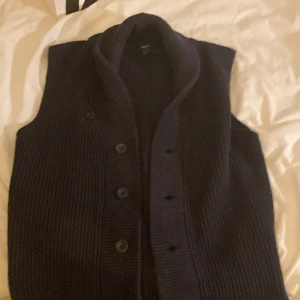 Mörkblå stickad väst från Filippa K - Snygg mörkblå stickad väst från Filippa K med bred sjalkrage och knappar framtill. Västen har en ribbstickad struktur som ger en cool och tidlös look. Perfekt att slänga över en skjorta eller t-shirt för extra stilpoäng.