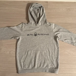 Grå hoodie från Sail Racing - Snygg grå hoodie från Sail Racing med svart logotyp tryckt på bröstet. Tröjan har huva, lång ärm och en klassisk magficka framtill. Perfekt för dig som gillar sportig och avslappnad stil. Materialet är mjukt och känns skönt mot huden.