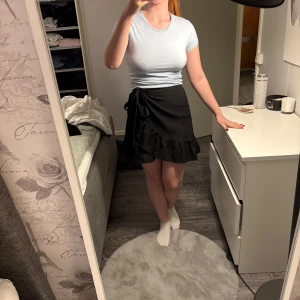 Svart omlottkjol med volang - Snygg svart omlottkjol med volangkant och knytning i sidan. Kjolen är kort och har en feminin och trendig look som passar perfekt till en enkel topp eller stickad tröja. Materialet känns lätt och följsamt.